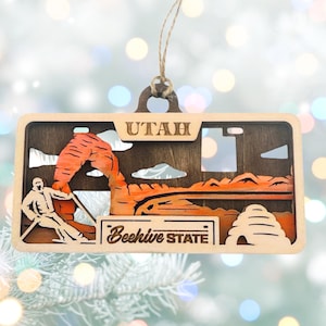 May include: Wooden Utah ornament featuring a layered design with the state name at the top. The ornament depicts a landscape with an arch, a person, and the words "Beehive State". The ornament is suspended by a natural twine.