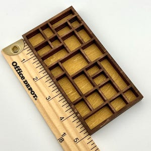 May include: A rectangular wooden shadow box display case with multiple compartments of varying sizes, featuring a dark brown frame and a light tan back panel. A wooden ruler is placed alongside, showing the object is approximately 5 inches long.