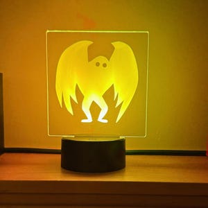 May include: A glowing yellow night light featuring a bat-like creature with outstretched wings. The light is square-shaped and sits on a black base, perfect for a Halloween or spooky-themed room.