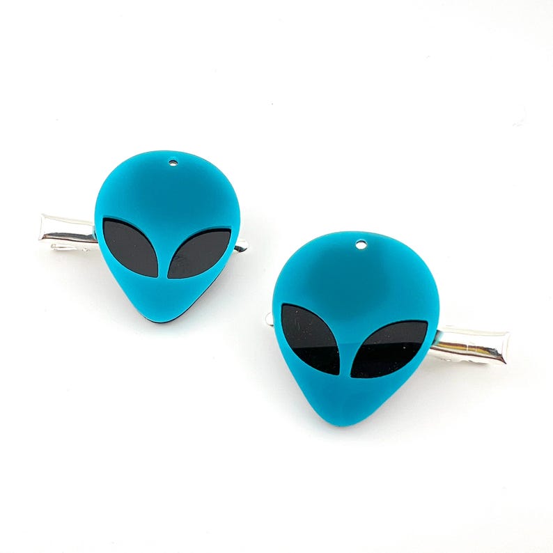 Op de afbeelding: Een paar teal blauwe alien hoofd haarclips met zwarte ogen. De clips zijn gemaakt van acryl en hebben een zilveren metalen balk.