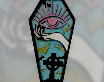 May include: A black coffin-shaped pin with a blue and green background. The pin features a white hand reaching out from the coffin, a pink eye with a sunburst, and a black Celtic cross. There are gold bats flying around the pin.