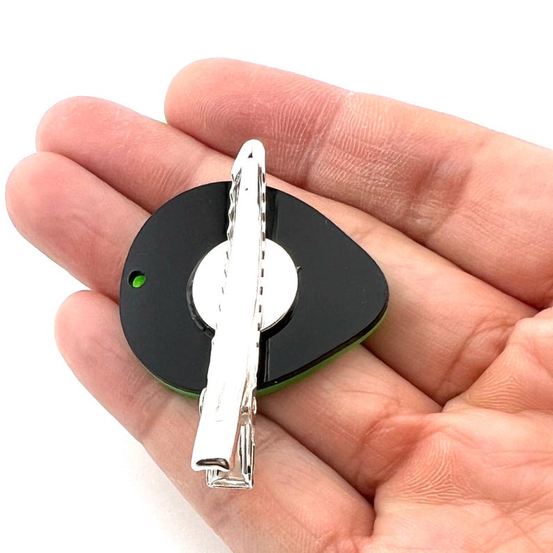 Op de afbeelding: Een zwarte en groene avocado-vormige haarspeld met een zilveren metalen sluiting. De speld wordt in een hand gehouden.