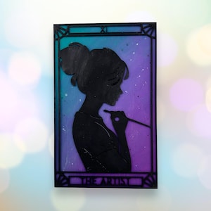 May include: A black silhouette of a person holding a paintbrush, set against a purple and blue gradient background. The image is framed in black and has the text "THE ARTIST" at the bottom.