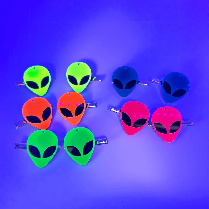 Op de afbeelding: Acht neonkleurige alien-hoofd haarclips in verschillende kleuren, waaronder groen, blauw, roze en oranje. De alien-hoofden hebben zwarte ogen en zijn bevestigd aan zilveren haarclips.