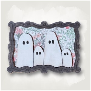 May include: A rectangular wall decor piece featuring four white wooden ghost cutouts with black painted eyes. The ghosts are set against a floral background with pink and green botanical patterns, all enclosed in a dark, ornate wooden frame.
