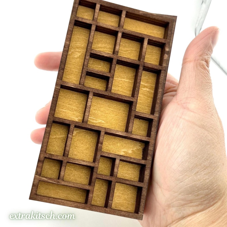May include: A rectangular wooden display tray featuring a grid of various sized compartments with dark brown edges and a light tan interior. The tray has a flat base and raised dividers, creating multiple open sections for small items.