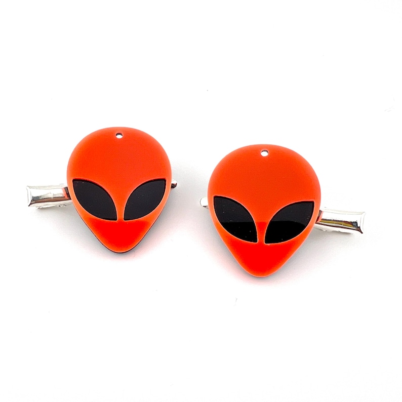 Op de afbeelding: Twee oranje haarclips met een alienhoofd en zwarte ogen. De clips zijn gemaakt van acryl en hebben een zilveren metalen clip.