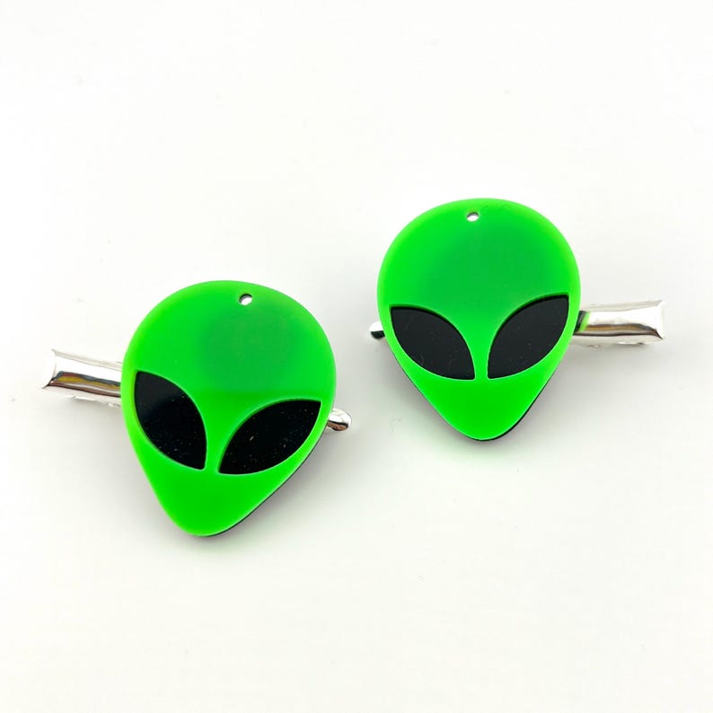 Op de afbeelding: Twee groene alien-hoofd haarclips met zwarte ogen. De clips zijn gemaakt van plastic en hebben een zilveren metalen staaf.