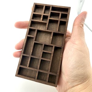May include: A rectangular dark brown wooden display shelf featuring a grid of multiple small, open compartments of varying sizes. The structure is composed of thin wooden dividers arranged in an asymmetrical pattern, creating a series of cubbies.