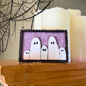 May include: A framed Halloween decoration featuring five white ghost figures with black eyes against a pink floral background. The frame is dark brown with a scalloped edge. A spider web is drawn in the upper left corner.