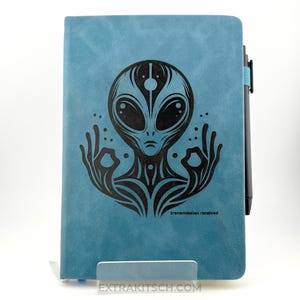 May include: A teal notebook with a black alien design on the cover. The alien has large eyes and is in a meditative pose. The text "transmission received" is printed at the bottom. A black pen is attached to the side.