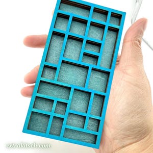 May include: A rectangular wooden grid organizer featuring a bright blue frame and a textured teal interior. The structure consists of multiple compartments of varying sizes arranged in an asymmetrical pattern. The text extrakitsch.com is visible.
