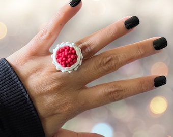 May include: A white ring with a red and white center, featuring a cluster of small red balls. The ring is worn on a person's finger.