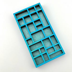 May include: A rectangular wooden tray featuring a bright turquoise painted frame with a grid of various sized compartments. The interior base of each section is lined with a textured, light teal fabric, creating a multi-level display surface.