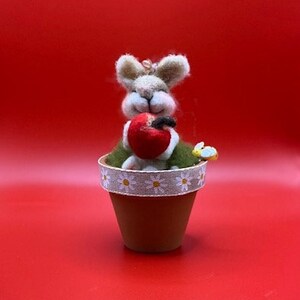 May include: A felt bunny holding a red apple sits in a small terracotta pot with a white daisy patterned ribbon around the rim.