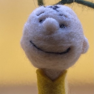 Peut inclure: Une décoration en feutre représentant un personnage de dessin animé souriant avec des yeux noirs et une chemise jaune. Le personnage est suspendu à une ficelle.