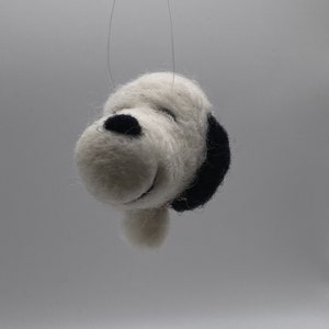 Peut inclure: Une décoration en feutre blanc représentant un chien avec des oreilles noires et un nez noir. La décoration est suspendue à une ficelle blanche.