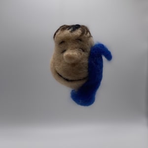 Peut inclure: Une figurine en feutre d'un personnage de dessin animé portant une écharpe bleue. Le personnage a une tête brune, un visage beige et un sourire aux yeux fermés.