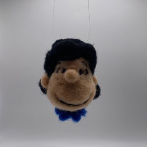 Peut inclure: Ornement en feutre représentant la tête d'un personnage de dessin animé avec des cheveux noirs, un visage beige et un nœud papillon bleu. L'ornement est suspendu par un fil transparent. Le personnage affiche un sourire.