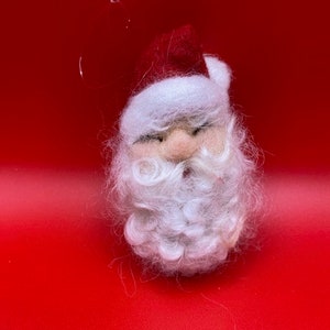 May include: A small felt Santa Claus ornament with a red hat, white beard, and a white fur trim. The ornament is against a red background.
