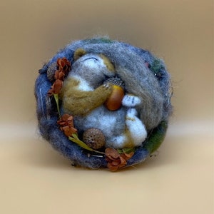 May include: A felted wool sculpture of a cat curled up in a nest of blue, green, and brown wool. The cat is holding a small acorn in its paws.