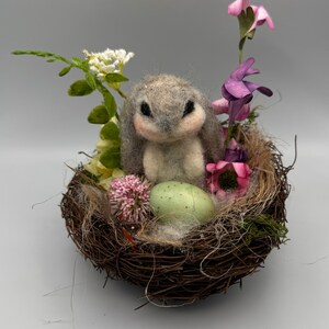 Könnte beinhalten: Handgefertigte Osterdekoration mit einem grauen Filzhasen in einem gewebten Nest. Das Nest ist mit künstlichen Blumen in Rosa, Lila und Weiß geschmückt und enthält ein hellgrünes, gesprenkeltes Ei. Der Hase hat rosa Akzente.