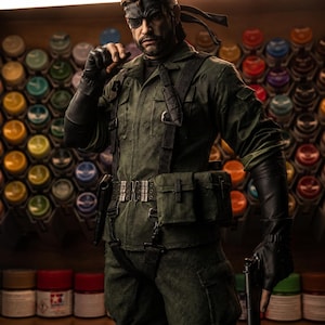 May include: A detailed figurine of a character in a green military-style uniform, black gloves, and an eye patch. The figure is holding a gun and has a serious expression. The background features a collection of paint bottles.