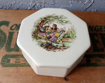 May include: White octagonal ceramic trinket box with a lid. The box features a colorful illustration of two people on a swing in a garden setting.