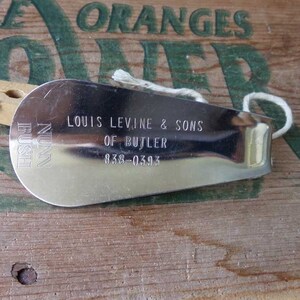 May include: A silver-colored shoehorn with a rounded end and a loop for hanging. The text "LOUIS LEVINE & SONS OF BUTLER 838-0393" is engraved on the surface. A wooden clothespin and a white string are also visible.