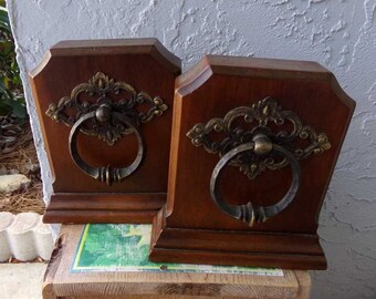 May include: A pair of wooden bookends with ornate metal handles. The handles are gold and have a ring shape. The bookends are a dark brown wood.