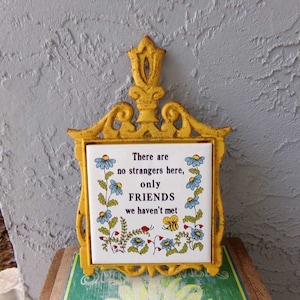 May include: Yellow cast iron hot pad with a white ceramic tile insert. The tile has a floral design and the text "There are no strangers here, only FRIENDS we haven't met."