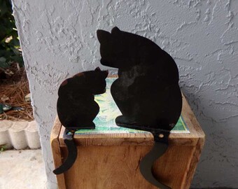 May include: Two black metal cat silhouettes, one larger than the other, are sitting side-by-side on a wooden box. The cats' tails are curled up behind them.