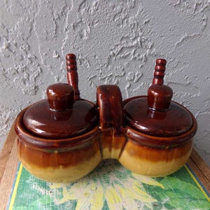May include: A brown and tan ceramic double-sided condiment holder with two lids and wooden handles. The holder is shaped like two connected pots.