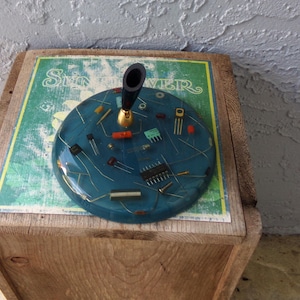 May include: A unique, handcrafted pen holder with a teal blue resin base, embedded with various electronic components. The pen holder features a black pen with a gold-colored base. The base rests on a vintage "Summer" sign.