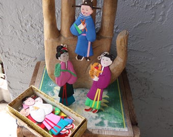 Pode incluir: Uma coleção de figuras de madeira planas e pintadas que representam pessoas com trajes tradicionais. Três figuras são exibidas na vertical, enquanto várias outras estão empilhadas ordenadamente dentro de uma caixa retangular com bordas douradas. Cada figura apresenta expressões faciais detalhadas e roupas coloridas.