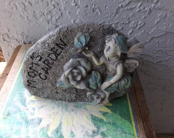 Pode incluir: Uma placa de jardim cinzenta com aspeto de pedra e o texto em relevo MOM'S GARDEN. A placa apresenta uma figura de fada esculpida com detalhes em azul-petróleo, asas e saia, estendendo a mão para uma borboleta azul. Duas rosas estão esculpidas.