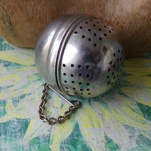 May include: A silver egg-shaped tea infuser with a chain handle. The infuser has a perforated design and is made of metal.