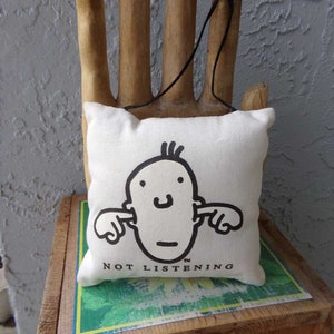 May include: A white canvas pillow with a black and white illustration of a person covering their ears with their hands. The pillow has the text "NOT LISTENING" printed on it.