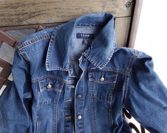 Pode incluir: Uma jaqueta jeans azul com gola clássica, mangas compridas e fecho frontal com botões. A peça inclui dois bolsos no peito com abas abotoadas, costuras na cor cobre e uma etiqueta Izod Jeans na parte interna da gola.