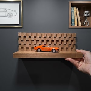 May include: A rectangular wooden floating shelf featuring a textured, grid-like back panel made of small, protruding wooden blocks. An orange die-cast toy car is positioned on the flat surface of the shelf, which has a natural wood grain finish.