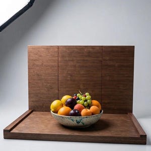May include: A ceramic bowl filled with a variety of fresh fruit, including green grapes, yellow lemons, oranges, and dark purple plums, resting on a dark brown wooden photography backdrop with a vertical panel and a flat base.