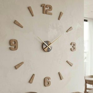 May include: A large wall clock featuring individual wooden numerals for 3, 6, 9, and 12, interspersed with wooden wedge-shaped markers. The center mechanism is a circular metallic disc with two thin, straight gold-toned hands indicating the time.