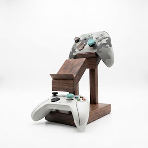 May include: A dark wood gaming controller stand featuring two tiers. The top tier holds a gray and white camouflage controller with light blue thumbsticks, while the bottom tier holds a solid white controller with light blue thumbsticks.