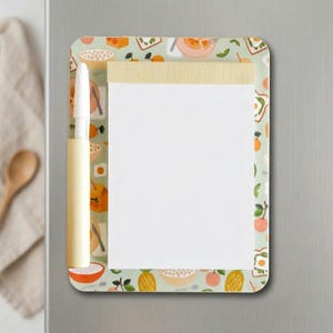 Può includere: Un blocco note magnetico rettangolare con bordo verde chiaro caratterizzato da un motivo colorato a tema colazione con ciotole, frutta e toast. Il blocco ha una sezione di carta bianca per scrivere e un portapenne color crema sul lato sinistro.