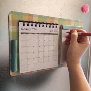 Peut inclure: Calendrier magnétique de janvier 2026 avec un bloc-notes attaché. Le calendrier est maintenu en place par un support à carreaux vert et jaune. Une personne écrit sur le bloc-notes avec un stylo rouge. Un aimant rose est visible.