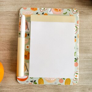 May include: A rectangular notepad with a light green border featuring a breakfast-themed pattern of oranges, bowls, and eggs. The notepad has a cream-colored top binding and a white paper surface, accompanied by a white pen held in a matching holder.