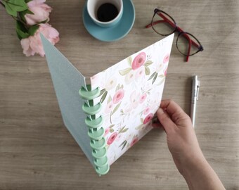 May include: A light blue notebook with a floral design on the cover. The notebook has a green spiral binding and is open to reveal a blank page.