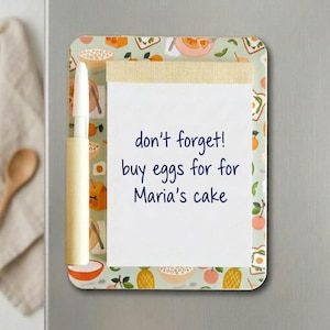 May include: A rectangular memo board with a notepad and pen. The board features a light green background with food illustrations. The notepad has the text "don't forget! buy eggs for Maria's cake".