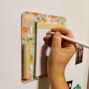 May include: A white dry-erase board with a light green and orange patterned frame. The frame has a small beige roll on the left side. A hand is writing on the board with a white pen.