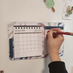 Può includere: Un set magnetico di calendario e blocco note su una superficie bianca. Il calendario mostra "Gennaio 2026" e una griglia mensile. Una persona scrive sul blocco note con una penna rossa. Il set ha un design arcobaleno pastello.